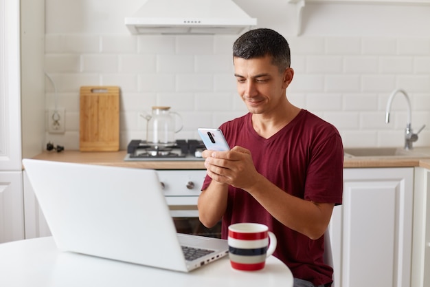 Hombre guapo positivo que trabaja en línea en casa en una computadora portátil, usando un teléfono inteligente para revisar correos electrónicos mientras tiene un descanso para el trabajo independiente, mirando la pantalla del dispositivo, escribiendo un mensaje.