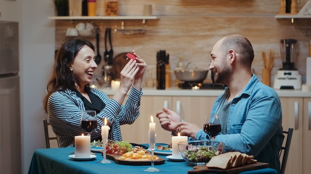 Hombre guapo proponiendo matrimonio a su novia durante la cena festiva, en la cocina sentado a la mesa bebiendo una copa de vino tinto. Feliz mujer sorprendida sonriendo y abrazándolo.