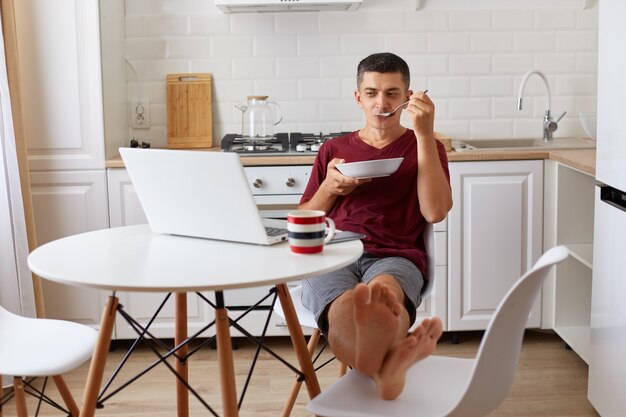 Hombre guapo relajado con camiseta informal granate sentado en la mesa, poniendo los pies en una silla, comiendo sopa y viendo una película, disfrutando del desayuno durante el fin de semana o mientras se toma un descanso del trabajo en línea.