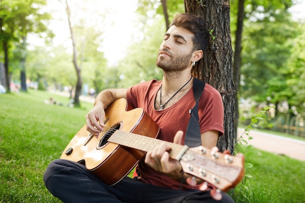 Hombre guapo romántico descansando en el parque con instrumento. Músico sentarse sobre el césped y tocar la guitarra