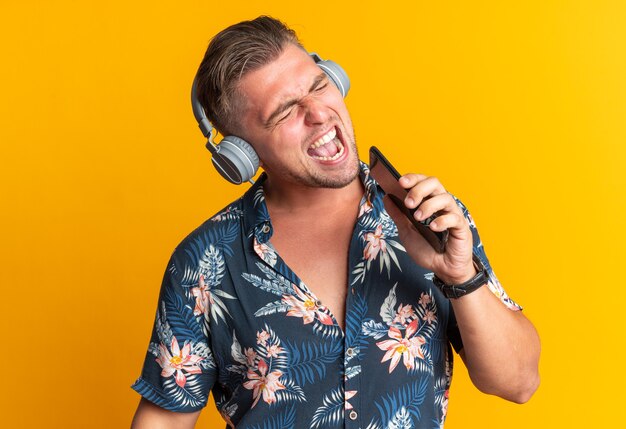Hombre guapo rubio alegre en auriculares sosteniendo el teléfono cerca de su boca cantando aislado en la pared naranja con espacio de copia