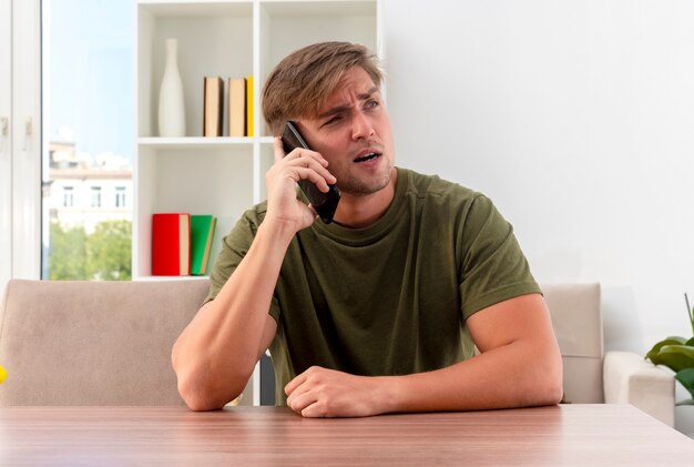 Hombre guapo rubio joven confiado se sienta a la mesa hablando por teléfono y mirando a un lado