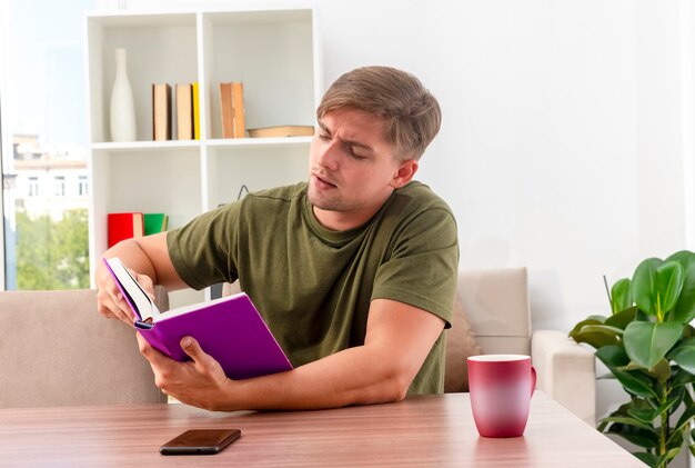 Hombre guapo rubio joven confiado se sienta a la mesa con la taza y el teléfono sosteniendo y mirando el libro dentro de la sala de estar