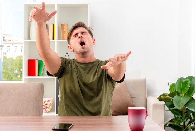 Hombre guapo rubio joven disgustado se sienta en la mesa levantando y sosteniendo las manos rectas con los ojos cerrados dentro de la sala de estar