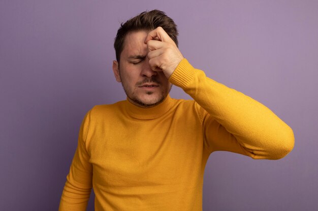 Hombre guapo rubio joven dolorido manteniendo la mano en la cabeza con los ojos cerrados aislado en la pared púrpura