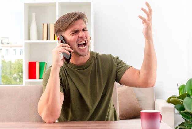 Hombre guapo rubio joven molesto se sienta a la mesa con la taza gritando a alguien en el teléfono con la mano levantada