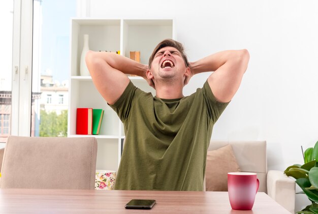 Hombre guapo rubio joven molesto se sienta a la mesa con la taza y el teléfono poniendo las manos en la cabeza detrás mirando hacia arriba