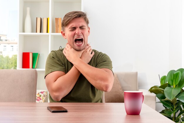 Hombre guapo rubio joven molesto se sienta a la mesa con el teléfono y la taza ahogándose con las dos manos dentro de la sala de estar