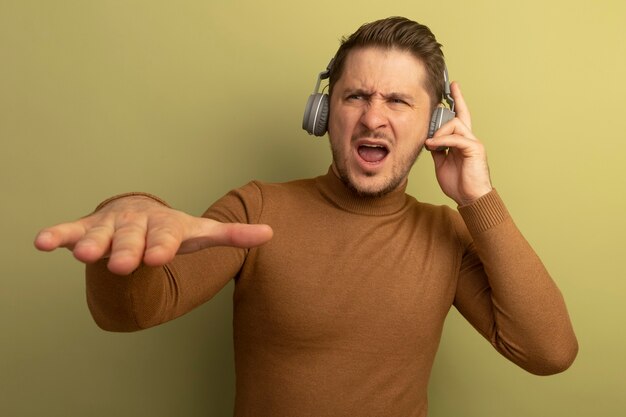 Hombre guapo rubio joven molesto usando y tocando los auriculares manteniendo la mano en el aire mirando de lado