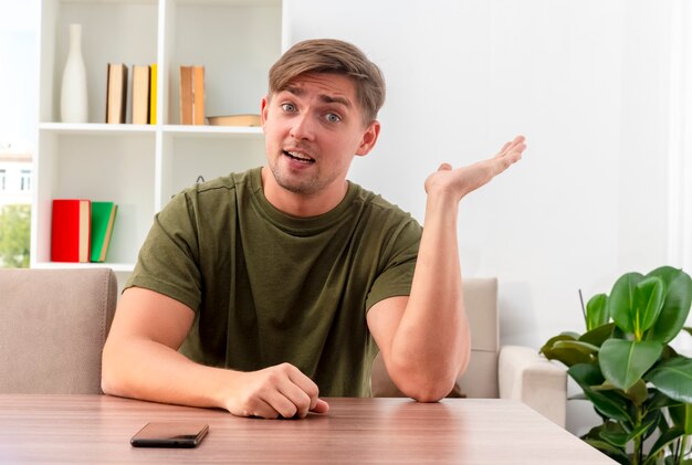 Hombre guapo rubio joven sorprendido se sienta a la mesa con el teléfono levantando la mano mirando a la cámara dentro de la sala de estar