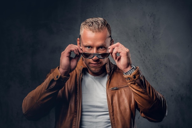 Hombre guapo, rubio y musculoso con gafas de sol, vestido con una camiseta blanca y una chaqueta de cuero marrón.