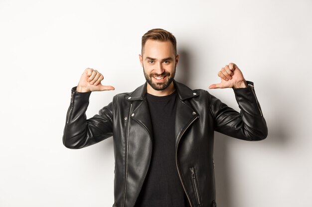 Hombre guapo seguro con chaqueta de cuero negro, apuntando a sí mismo y sonriendo seguro de sí mismo, de pie