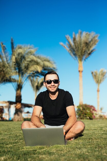 Hombre guapo sentado en la hierba y usando la computadora portátil.