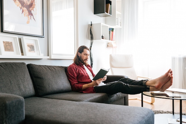 Hombre guapo sentado y leyendo el libro en el sofá en casa