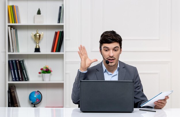 Hombre guapo de servicio al cliente en traje gris con computadora y auriculares hablando con el cliente