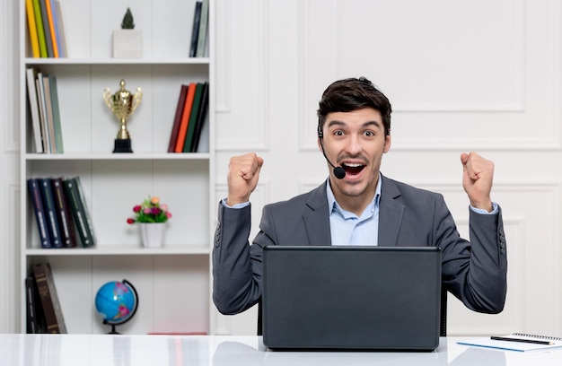 Hombre guapo de servicio al cliente en traje gris con computadora y auriculares levantando los puños emocionantemente