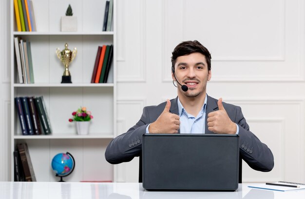 Hombre guapo de servicio al cliente en traje gris con computadora y auriculares que muestran un buen gesto