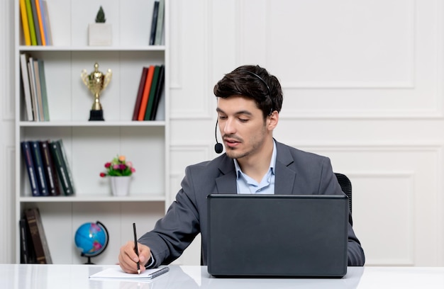 Hombre guapo de servicio al cliente en traje gris con computadora y auriculares tomando notas