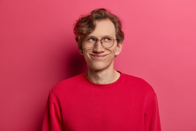 Hombre guapo soñador con expresión complacida, enfocado a un lado con expresión encantada, sonrisa tierna, imagina la primera cita, usa anteojos redondos y jersey rojo, tiene mirada feliz, aislado en rosa