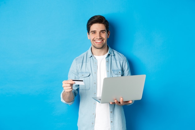 Hombre guapo sonriendo, comprando en línea y pagando por el producto, sosteniendo la tarjeta de crédito con el portátil, de pie sobre fondo azul.