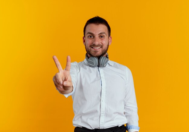 Hombre guapo sonriente con auriculares en el cuello gestos victoria signo de mano aislado en la pared naranja