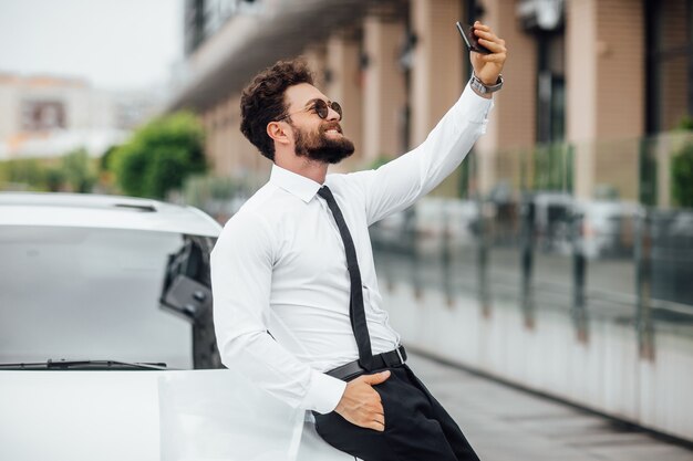 Hombre guapo, sonriente, barbudo con camisa blanca, haciendo selfie cerca de su nuevo coche al aire libre en las calles de la ciudad