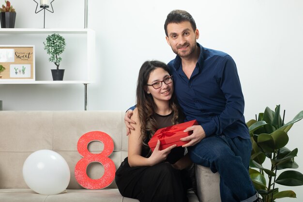 Hombre guapo sonriente dando caja de regalo roja a una mujer joven y bonita con gafas ópticas sentado en el sofá en la sala de estar en el día internacional de la mujer de marzo