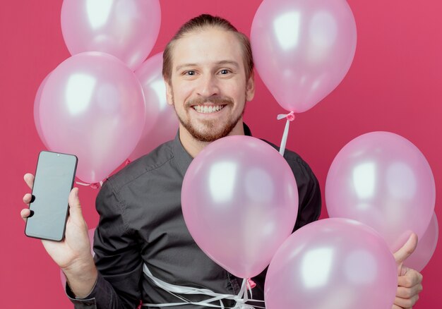 Hombre guapo sonriente se encuentra con globos de helio sosteniendo teléfono aislado