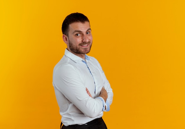 Hombre guapo sonriente se encuentra de lado cruzando los brazos aislado en la pared naranja