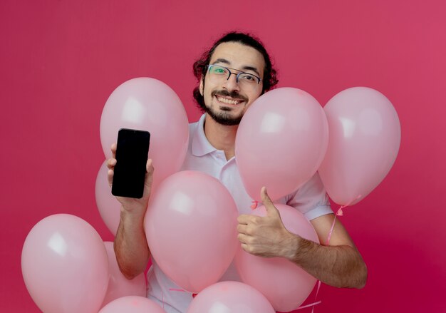 Hombre guapo sonriente con gafas de pie detrás de globos sosteniendo el teléfono con el pulgar hacia arriba aislado en la pared rosa