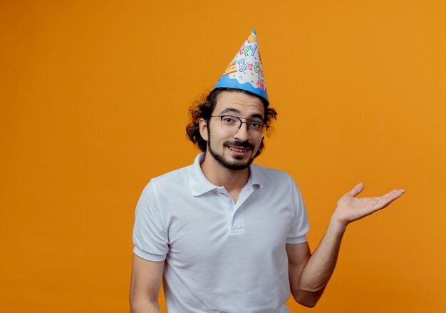 Hombre guapo sonriente con gafas y puntos de gorro de cumpleaños con la mano al lado aislado sobre fondo naranja con espacio de copia