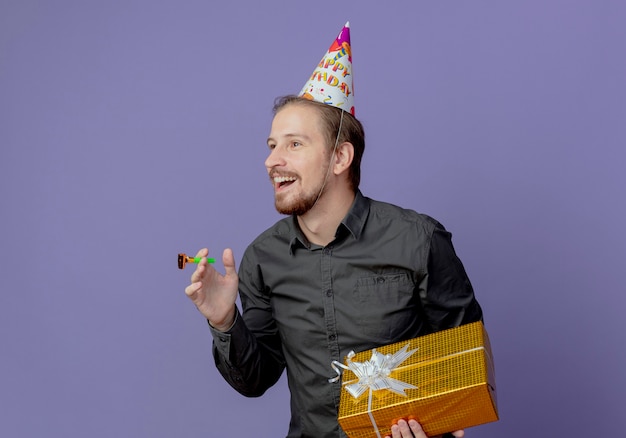 Hombre guapo sonriente en gorro de cumpleaños tiene caja de regalo y silbato mirando al lado aislado en la pared púrpura