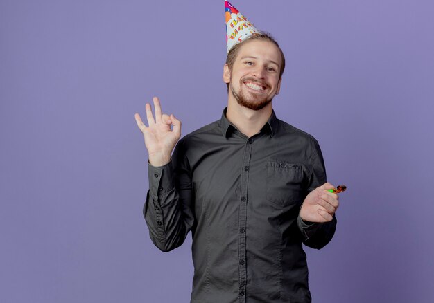 Hombre guapo sonriente en gorro de cumpleaños tiene silbato y gestos ok signo de mano aislado en la pared púrpura