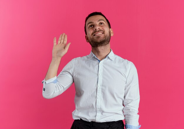 Hombre guapo sonriente levanta la mano mirando hacia arriba aislado en la pared rosa