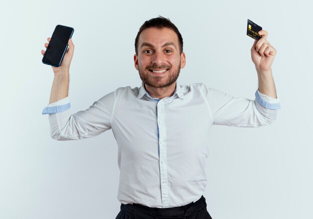 Hombre guapo sonriente levanta las manos sosteniendo el teléfono y la tarjeta de crédito aislado en la pared blanca