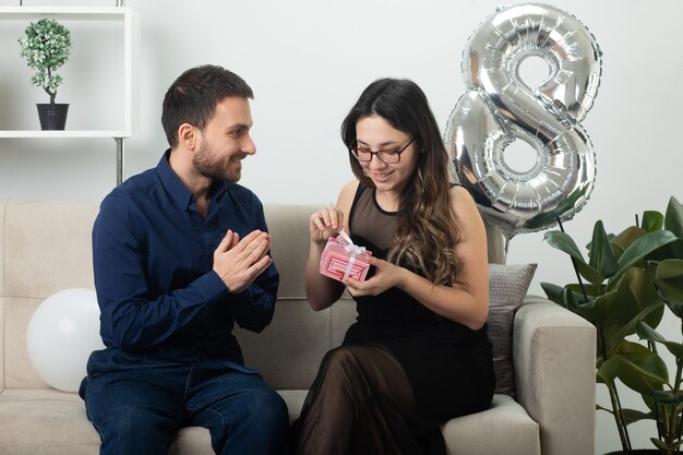 Hombre guapo sonriente mirando a una mujer joven y bonita complacida en gafas ópticas abriendo caja de regalo sentado en el sofá en la sala de estar en marzo día internacional de la mujer