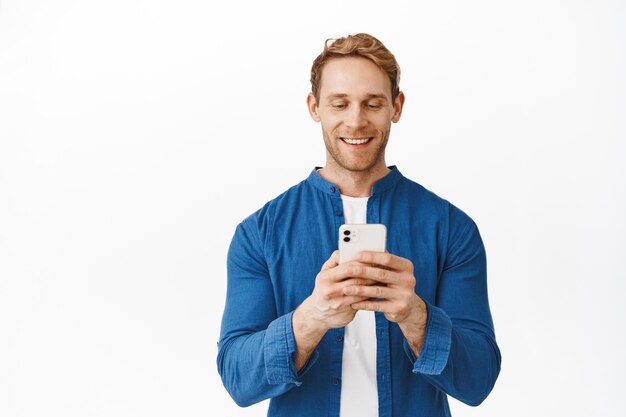 Hombre guapo sonriente mirando la pantalla de su teléfono, leyendo o viendo videos en la aplicación de teléfono inteligente, mirando complacido a la pantalla, tomando fotografías, de pie contra el fondo blanco