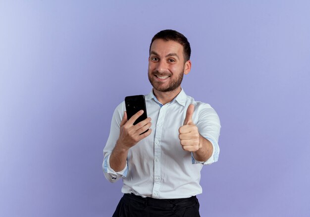 Hombre guapo sonriente pulgar hacia arriba y sostiene el teléfono aislado en la pared púrpura