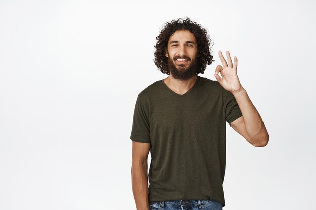 Un hombre guapo sonriente que muestra un signo de aprobación aprobado y como algo alabado es algo bueno de pie sobre un fondo blanco