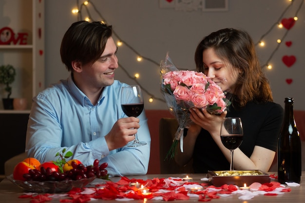 Hombre guapo sonriente sosteniendo una copa de vino y mirando a una mujer bonita oliendo el ramo de flores sentado a la mesa en la sala de estar en el día de San Valentín