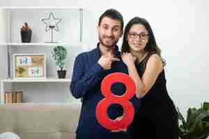 Foto gratuita hombre guapo sonriente sosteniendo la figura de ocho rojos y apuntando a una mujer joven y bonita con gafas ópticas de pie en la sala de estar en el día internacional de la mujer de marzo