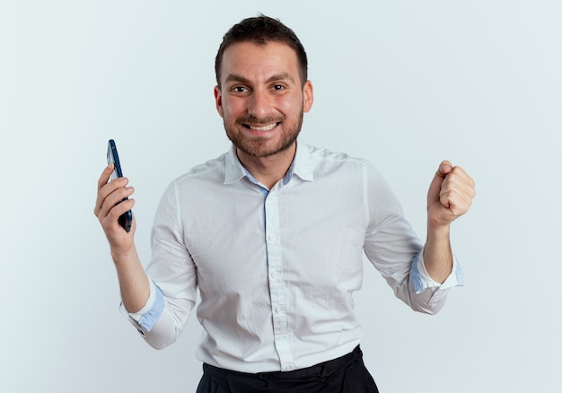 Hombre guapo sonriente sostiene el teléfono y mantiene el puño aislado en la pared blanca