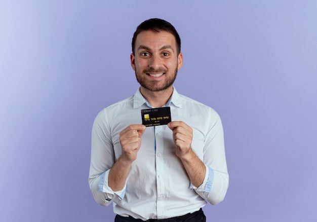 Hombre guapo sonriente tiene tarjeta de crédito mirando aislado en la pared púrpura