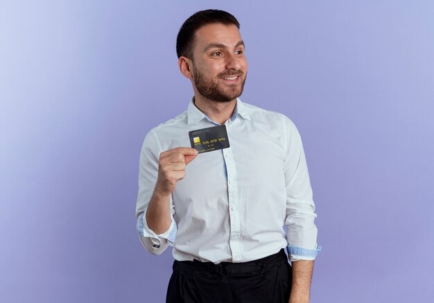 Hombre guapo sonriente tiene tarjeta de crédito mirando al lado aislado en la pared púrpura