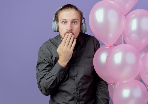 Hombre guapo sorprendido en auriculares se encuentra con globos de helio poniendo la mano en la boca aislada en la pared púrpura
