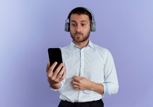 Hombre guapo sorprendido en auriculares sostiene y mira el teléfono aislado en la pared púrpura