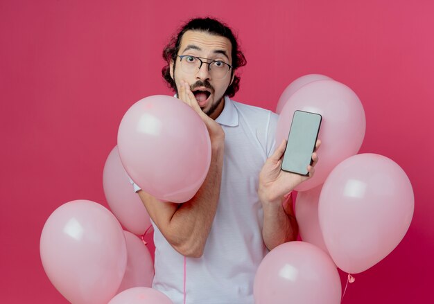 Hombre guapo sorprendido con gafas de pie entre globos sosteniendo el teléfono y poniendo la mano en la mejilla aislada en la pared rosa