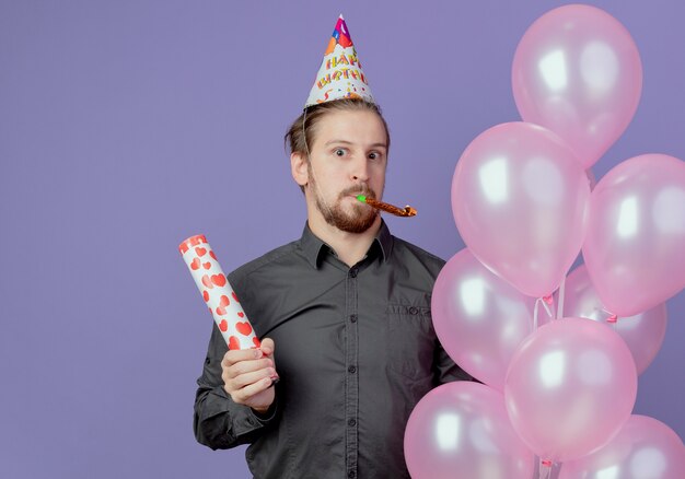 Hombre guapo sorprendido en gorra de cumpleaños sostiene globos de helio y cañón de confeti que sopla silbato aislado en la pared púrpura