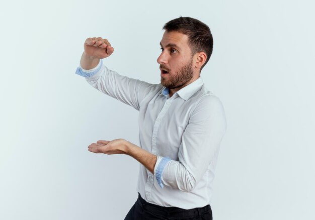 Hombre guapo sorprendido se para de lado pretendiendo sostener algo aislado en la pared blanca