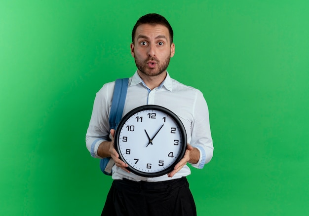 Hombre guapo sorprendido con mochila tiene reloj aislado en la pared verde